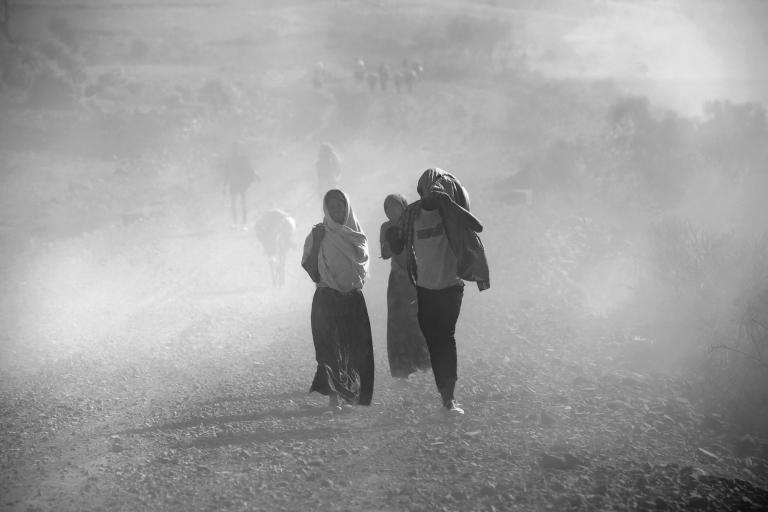 People walk along a dusty, rocky path in a barren landscape, partially shielding themselves from the blowing dust and haze.