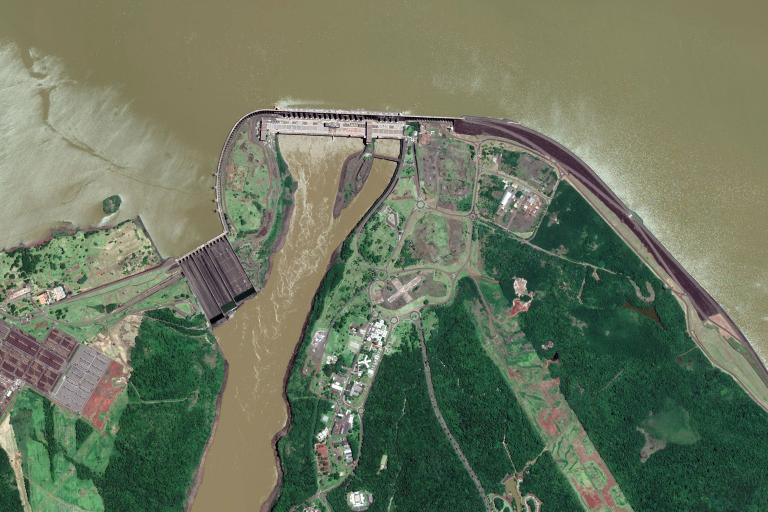 Satellite view of a large dam spanning a river with adjacent buildings, green vegetation, and brown water flowing through the structure.