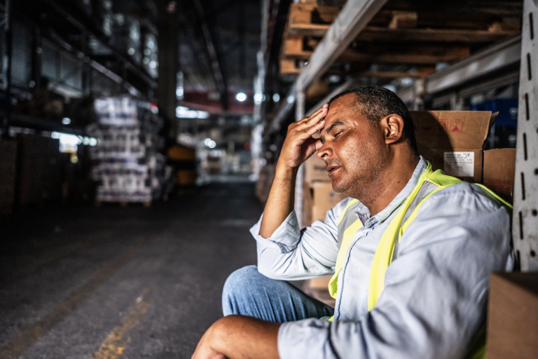A man in a high-visibility vest sits in a warehouse aisle, resting his head on his hand and appearing stressed or tired.