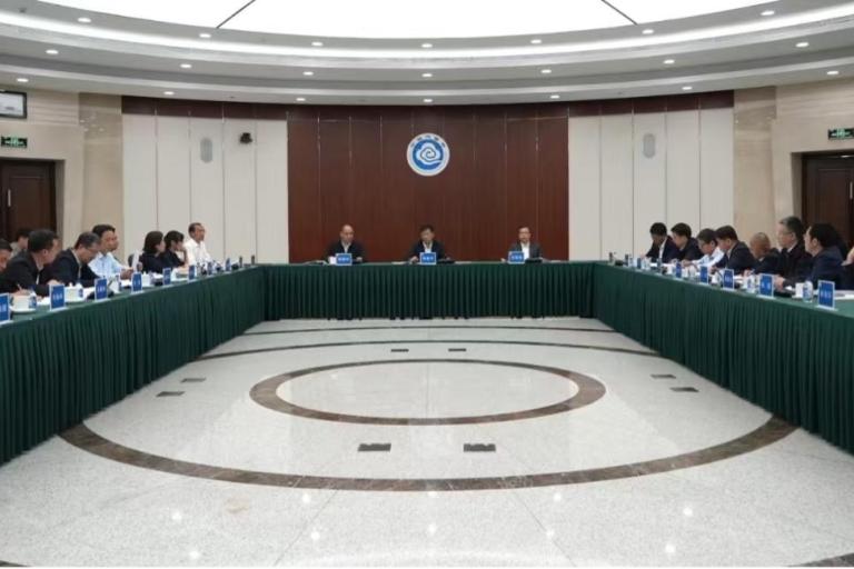 A formal meeting with over twenty people seated around a large U-shaped table in a conference room, with microphones and nameplates arranged in front of them.