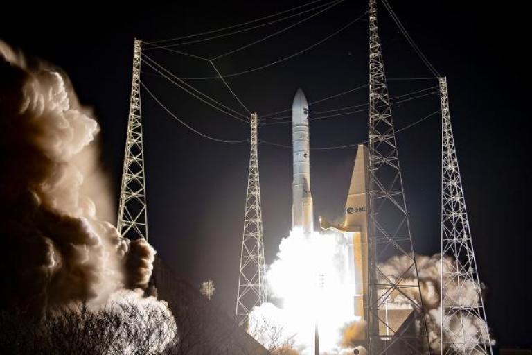 A rocket launches at night, producing bright flames and smoke, surrounded by tall support towers against a dark sky.