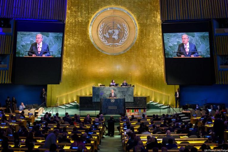 A speaker addresses delegates at the United Nations General Assembly hall, with two screens displaying his image and the UN emblem visible in the background.