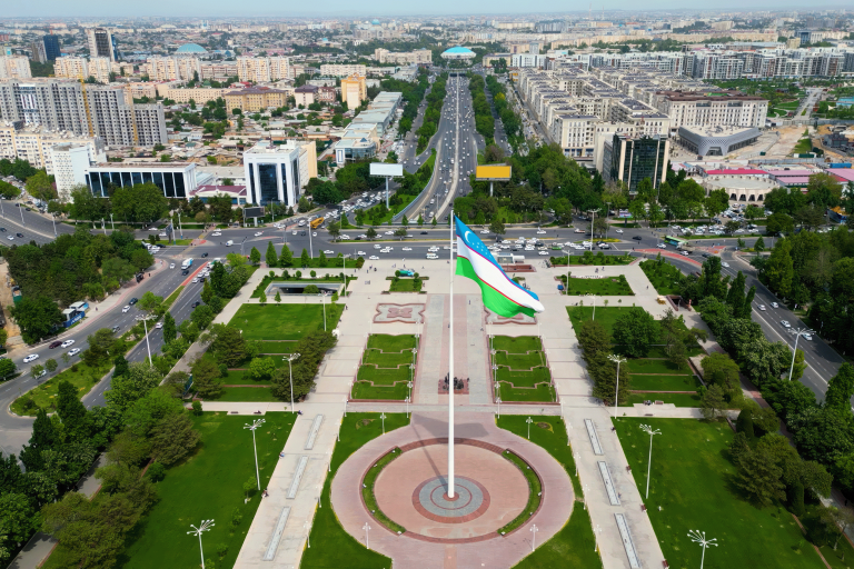 Aerial view of a city square with a large flagpole displaying the Uzbekistan flag, surrounded by green lawns, roads, and buildings.