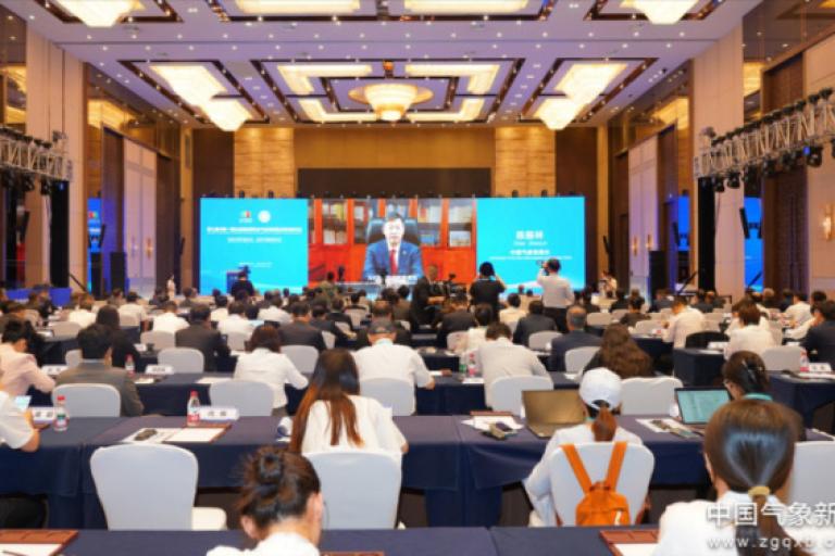 A large conference room filled with attendees facing a stage and screens displaying a speaker’s image during a formal event.
