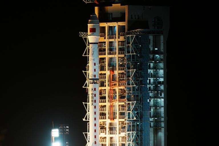 A rocket launches at night from a lit-up launch pad, emitting bright flames and smoke at the base against a dark sky.