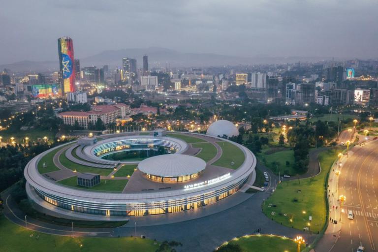 An aerial view of a modern, circular building with a central courtyard, surrounded by greenery and city lights, with a city skyline and tall buildings in the background at dusk.