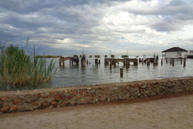A calm lake scene under a cloudy sky, showing partially submerged stone pillars and vegetation near the shore. A low stone wall runs along the sandy foreground, and a small white building with a dark roof stands on stilts over the water in the distance.