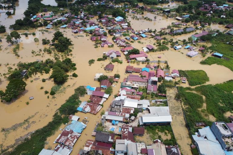 Aerial view of a flooded town with numerous buildings partially submerged in muddy water and surrounding areas also impacted by the flood.