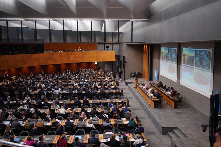 Большая группа людей принимает участие в официальной встрече в современном конференц-зале, где участники дискуссии сидят впереди, а слайды презентации проецируются на стену.