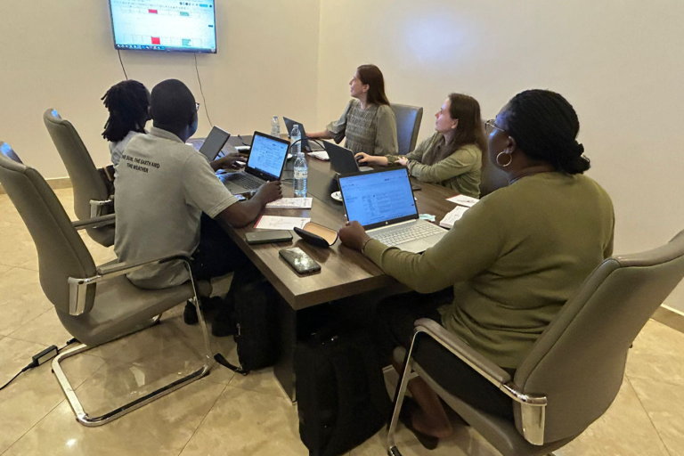 Five people sitting around a conference table with laptops, viewing a large screen displaying a spreadsheet in a meeting room.
