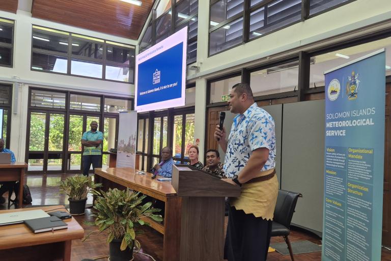 A man speaks at a podium during an indoor event, with attendees seated at tables and banners displaying "Solomon Islands Meteorological Service" visible in the background.