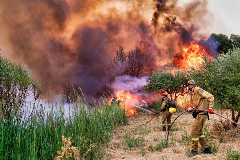Two firefighters in protective gear use hoses to battle a large wildfire, with thick smoke and tall flames consuming vegetation around them.