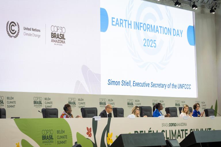 Panelists sit on stage at the COP30 Belém Climate Summit, with a large screen displaying "Earth Information Day 2025" and the name Simon Stiell, Executive Secretary of the UNFCCC.