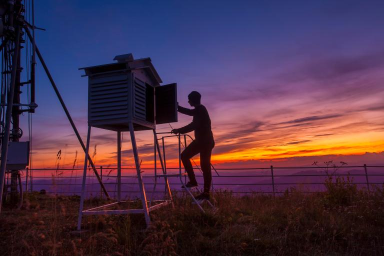 A person checks equipment inside a weather station at sunset, standing on a small ladder beside a fenced area and tall antenna structure.