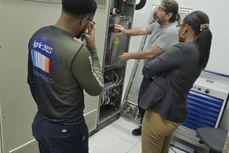 Three people stand in front of open server equipment in a lab, discussing the components; a laptop and documents are on a nearby table.