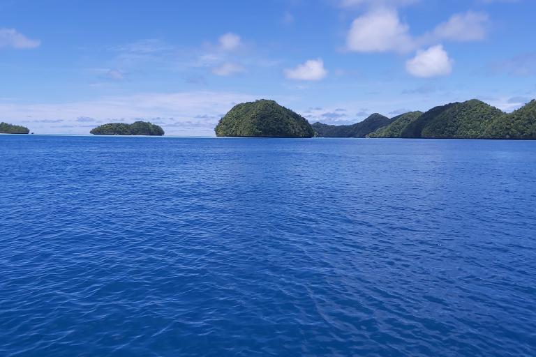 A calm, bright blue ocean with several small, green, forested islands under a partly cloudy sky.