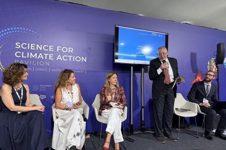 Five panelists sit and stand on a stage at the Science for Climate Action Pavilion, with a man speaking into a microphone and a presentation screen behind them.