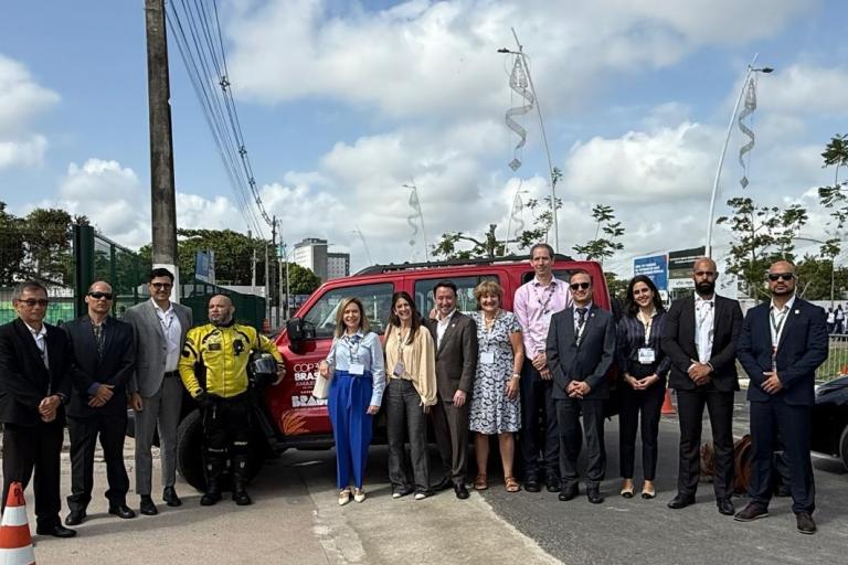 A group of people in business attire stand in a line outdoors in front of a red vehicle, with trees and a partly cloudy sky in the background.