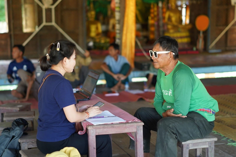 A woman interviews an older man in sunglasses indoors; she holds a phone and notebook, while others sit in the background.