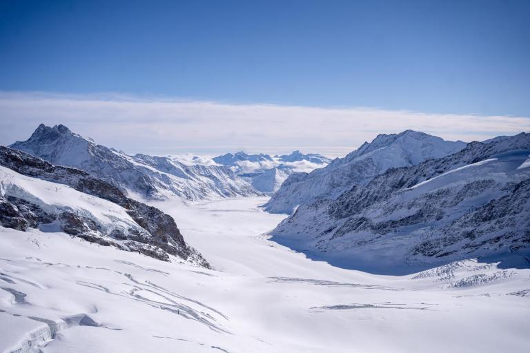 Snow-covered mountains and a glacier under a clear blue sky, with rugged peaks and a wide valley extending into the distance.