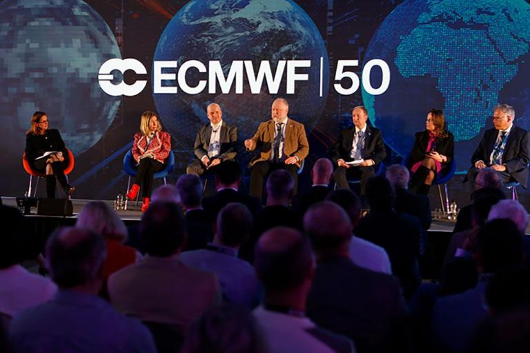 A panel of six people speaks on stage at an ECMWF 50th anniversary event, with an audience seated in front and large screens showing Earth in the background.