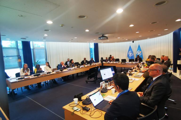 A group of people sit around a large conference table with laptops and documents in a meeting room, featuring United Nations flags in the background.