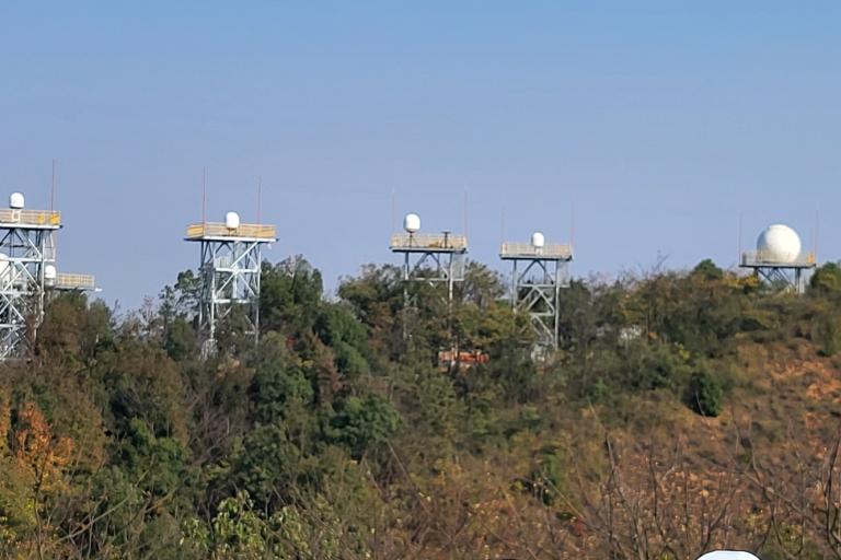 Several radar domes are mounted on tall metal towers above a forested hillside under a clear blue sky.