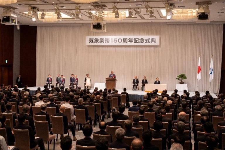 A formal ceremony in a large hall with seated attendees, a speaker at the podium, and banners in Japanese, alongside the Japanese flag and another flag.