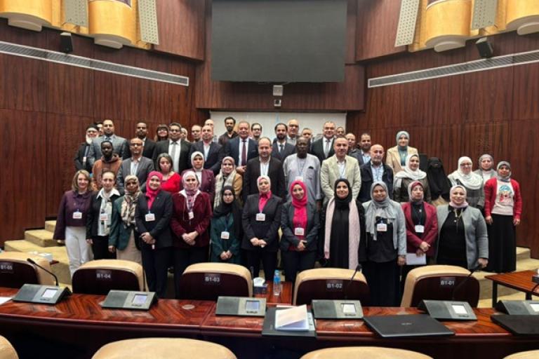A group of people pose for a group photo inside a large conference room with wooden paneling and empty seats in the foreground.