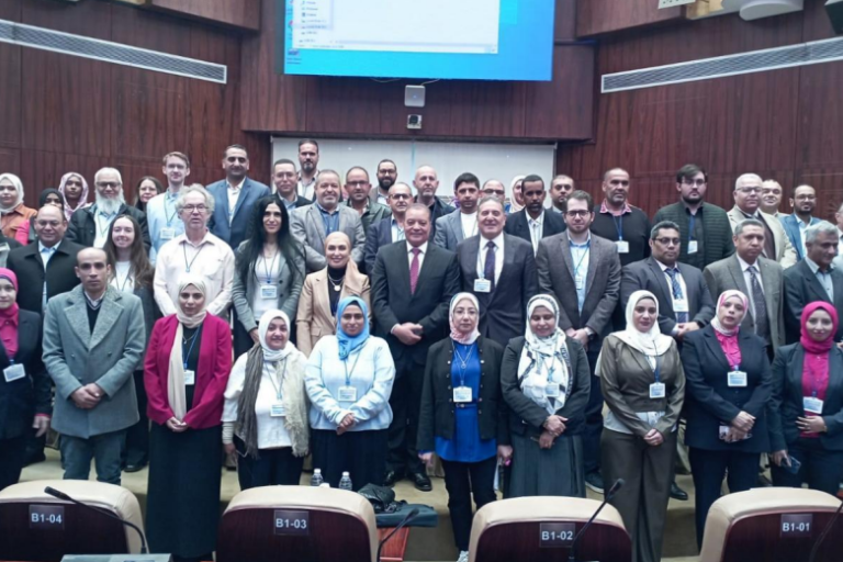 A large group of professionals, both men and women, pose together for a group photo in a conference room with a large screen behind them.