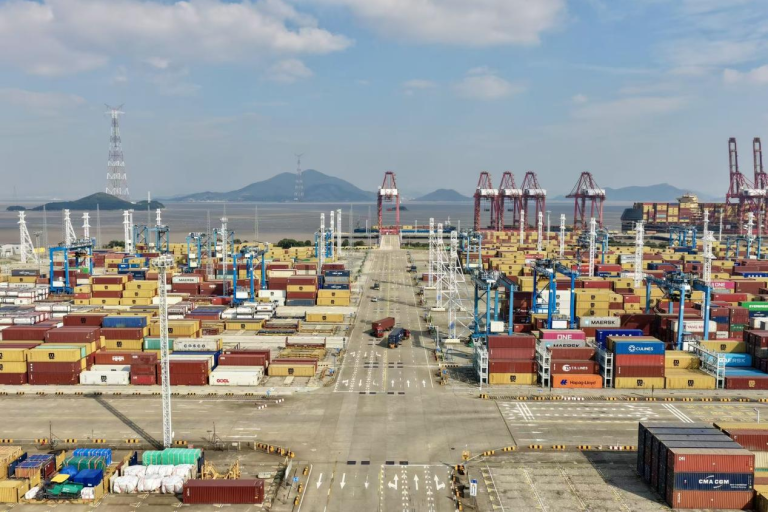 Aerial view of a large shipping port with stacked cargo containers, cranes, trucks, and distant water and hills under a partly cloudy sky.
