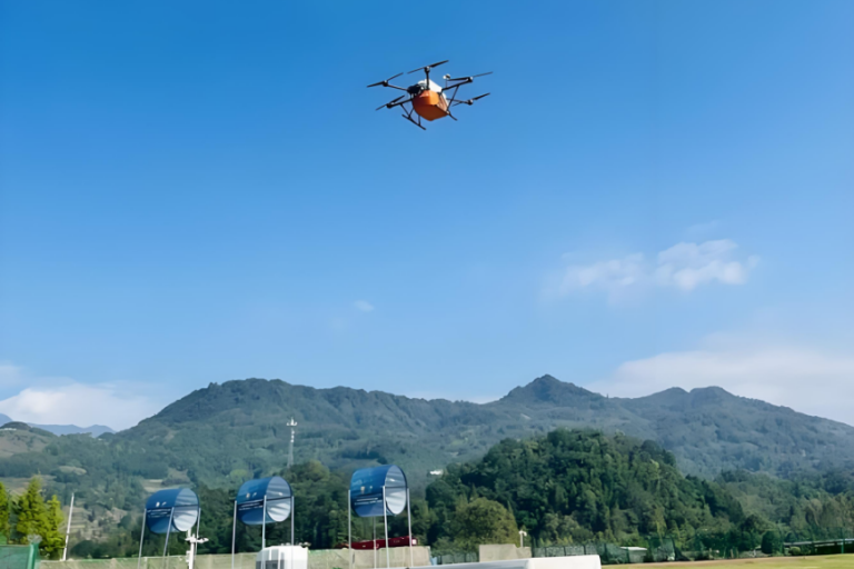 An orange drone with multiple rotors flies over a grassy field with distant hills and blue sky in the background.