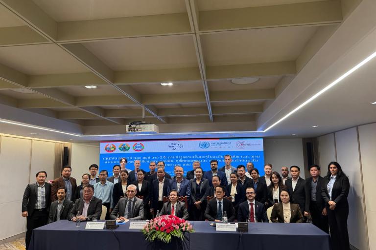 A group of people in business attire pose for a photo at a conference table, with a banner and flower arrangement in the background.