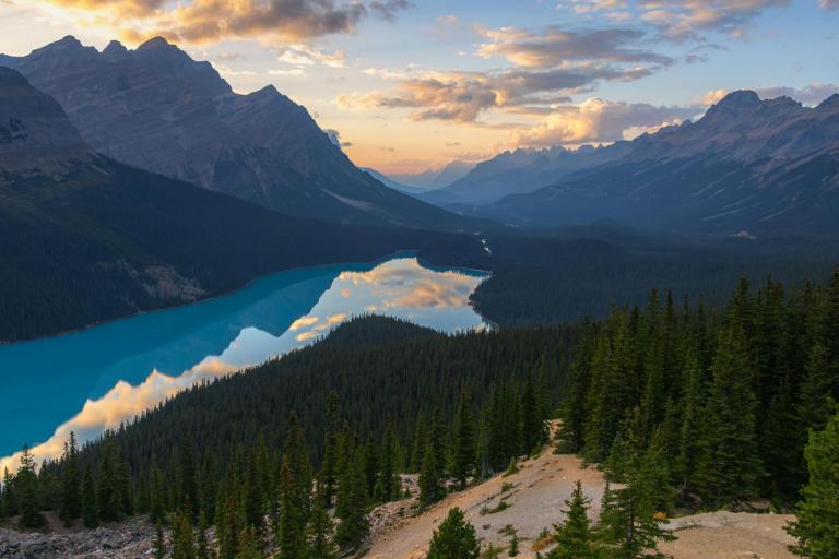 A turquoise lake winds through a forested valley surrounded by mountains at sunset, with clouds and sky reflected on the water's surface.