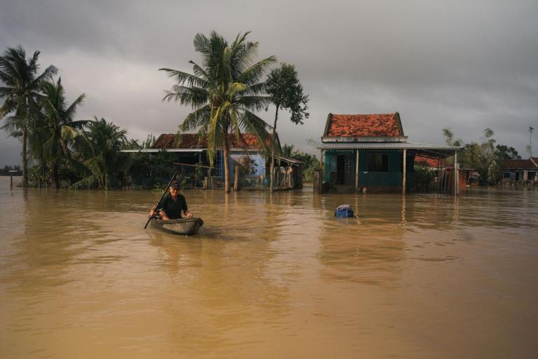 A person paddles a small boat through floodwaters in front of partially submerged homes and palm trees under a cloudy sky.