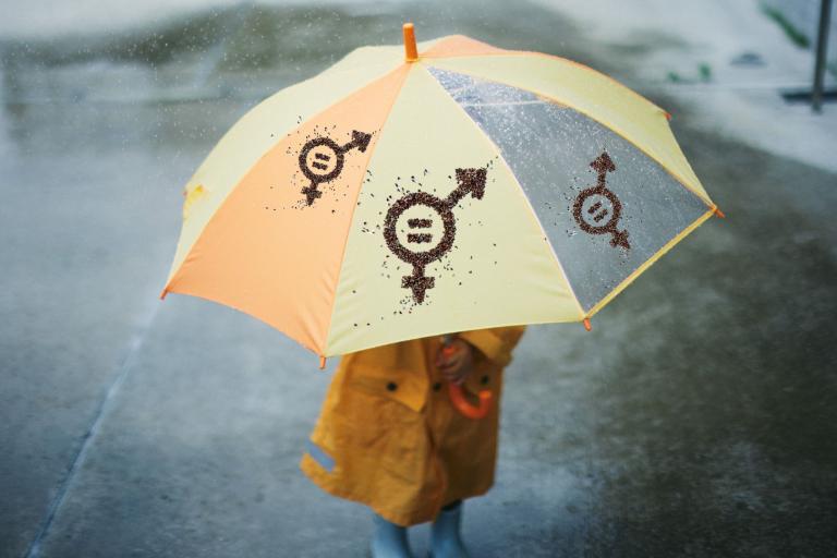 A child in a yellow raincoat stands under a yellow and orange umbrella with gender symbol designs on a rainy day.