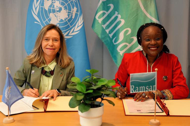 Two women sit at a table signing documents, with United Nations and Ramsar Convention flags in the background and a potted plant in the foreground.