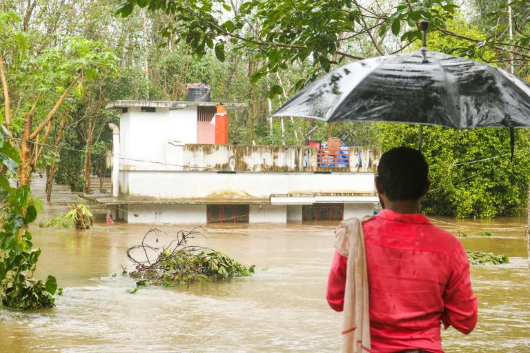 A person holding an umbrella observes a partially submerged house surrounded by floodwater and trees.
