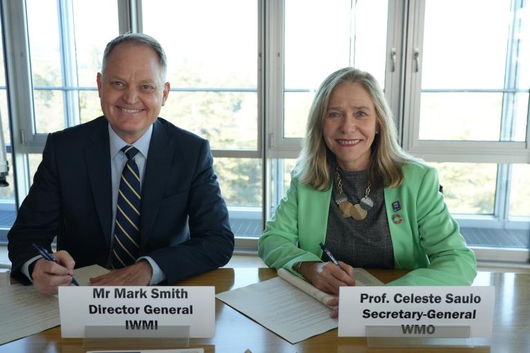 Two people sit at a table, each holding a pen and papers, with nameplates reading "Mr Mark Smith Director General IWMI" and "Prof. Celeste Saulo Secretary-General WMO.