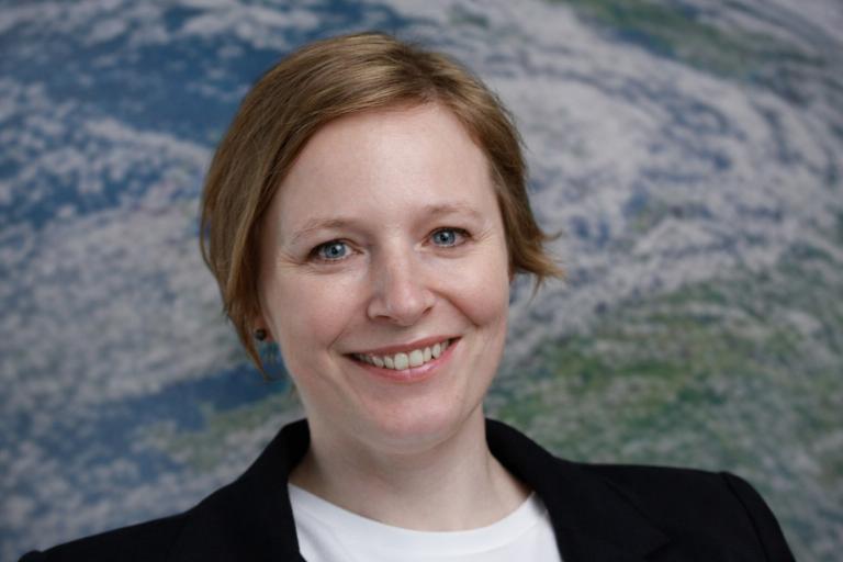 A woman with short light brown hair smiles at the camera, wearing a black blazer and white shirt, with a blurred Earth image in the background.