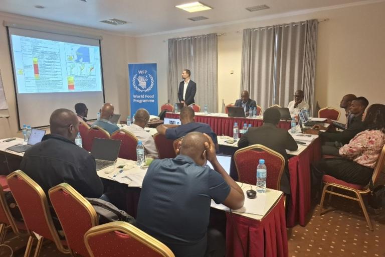 A group of people attend a presentation in a conference room with a World Food Programme banner and projected slides on the wall.
