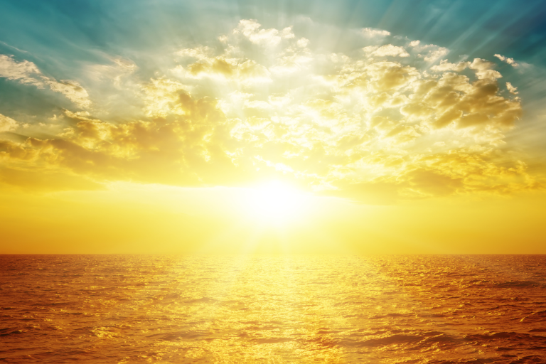 A bright golden sun rises or sets over the ocean, illuminating clouds and reflecting light on the water under a partly cloudy sky.