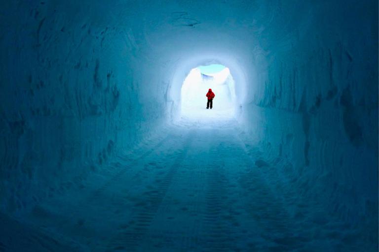 A person wearing a red jacket stands at the end of a tunnel made of snow and ice, illuminated by natural light.