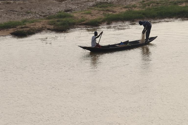 Two people in a wooden boat are fishing with a net on a calm river near a grassy and sandy riverbank.