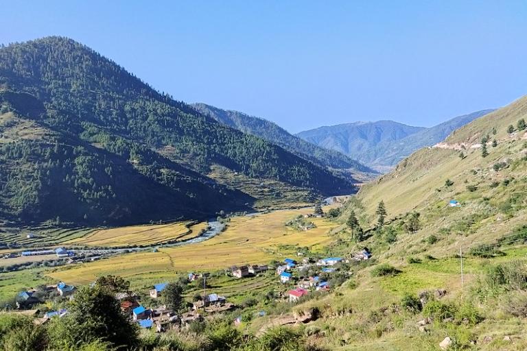 A river flows through a valley with yellow fields, small houses, and green hills under a clear blue sky.