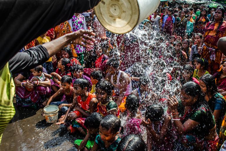 A group of children sit closely together as water is poured over them from a bucket, surrounded by a crowd of onlookers in colorful clothing.