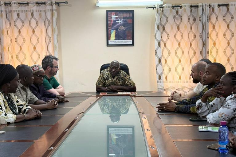 A group of people sit around a conference table, listening to a man seated at the head of the table in an office setting.
