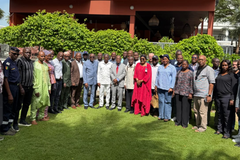A large group of adults stands together outside on a lawn in front of a building and greenery, posing for a group photo in daylight.