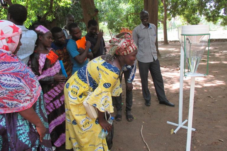 A group of people observes a demonstration of a water collection device outdoors under trees, with a woman in the foreground leaning in for a closer look.