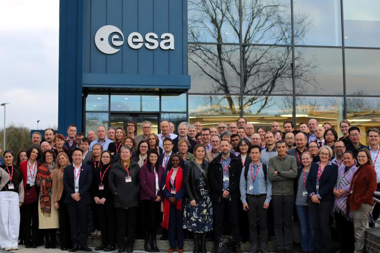 A large group of people stands in front of the ESA building, posing for a group photo. The glass facade reflects trees and sky in the background.